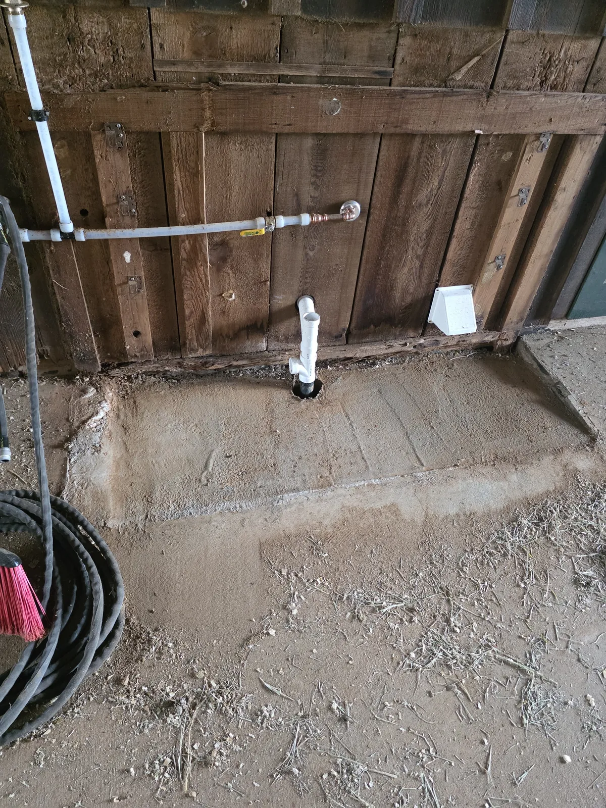 Barn floor patched in concrete around a PVC drain stub along a wooden wall