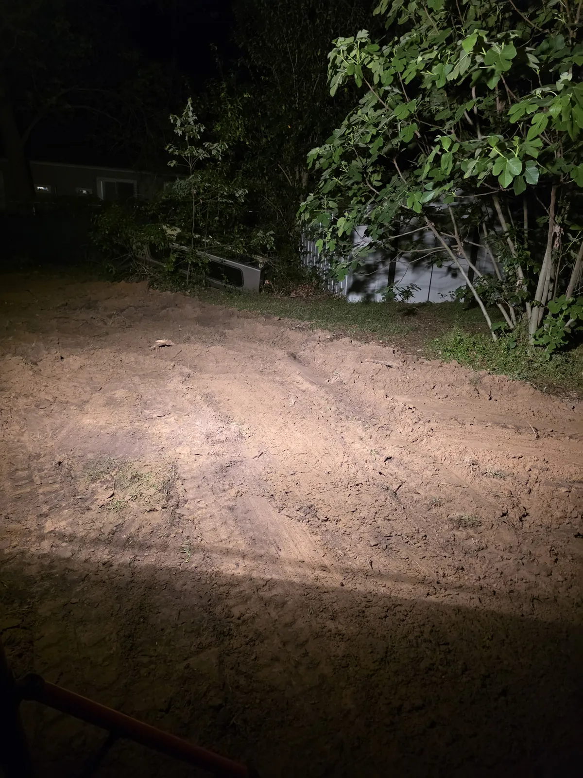 Leveled bare dirt where a trench was filled, lit by headlights beside shrubs at night